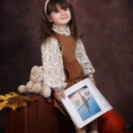 Уютные сказки осени: детская фотосессия в винтажном стиле Девочка показывает большую книгу с картинками, сидя на чемодане рядом с плюшевым зайцем и тыквой.