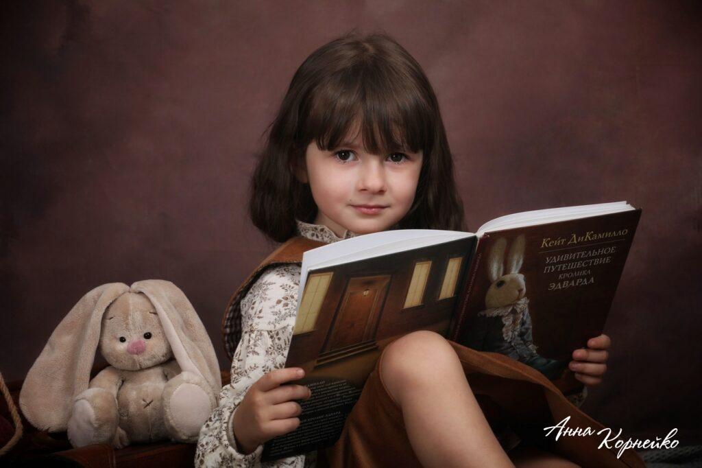 Крупный план девочки, читающей книгу Удивительное путешествие кролика Эдварда, рядом сидит бежевый плюшевый кролик.