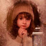 Девочка в вязаной шапке смотрит через заснеженное окно, домашняя фотосессия.